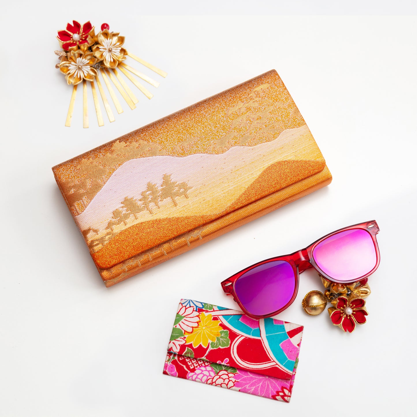 Bronze gold silk envelope clutch, sunglasses with floral card case, and decorative hairpin on a white background