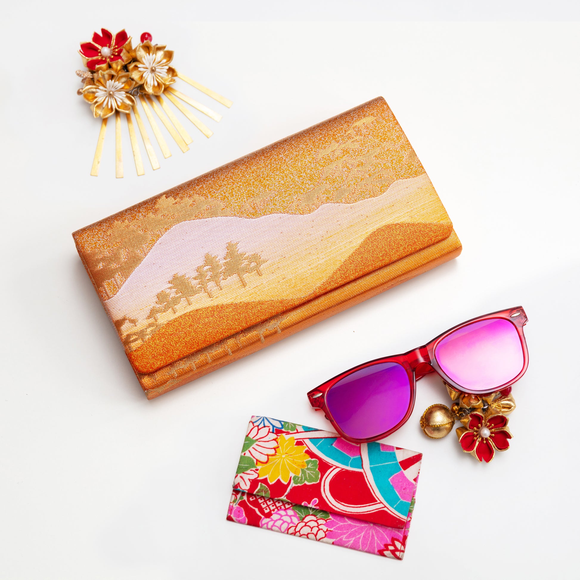 Bronze gold silk envelope clutch, sunglasses with floral card case, and decorative hairpin on a white background