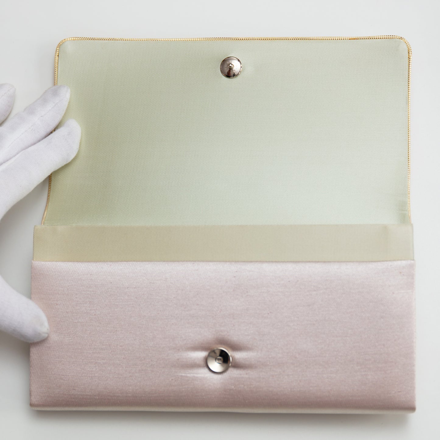 Pink metallic silk envelope clutch held open by gloved hand to show cream silk lining and silver metal stud fastener on white background.