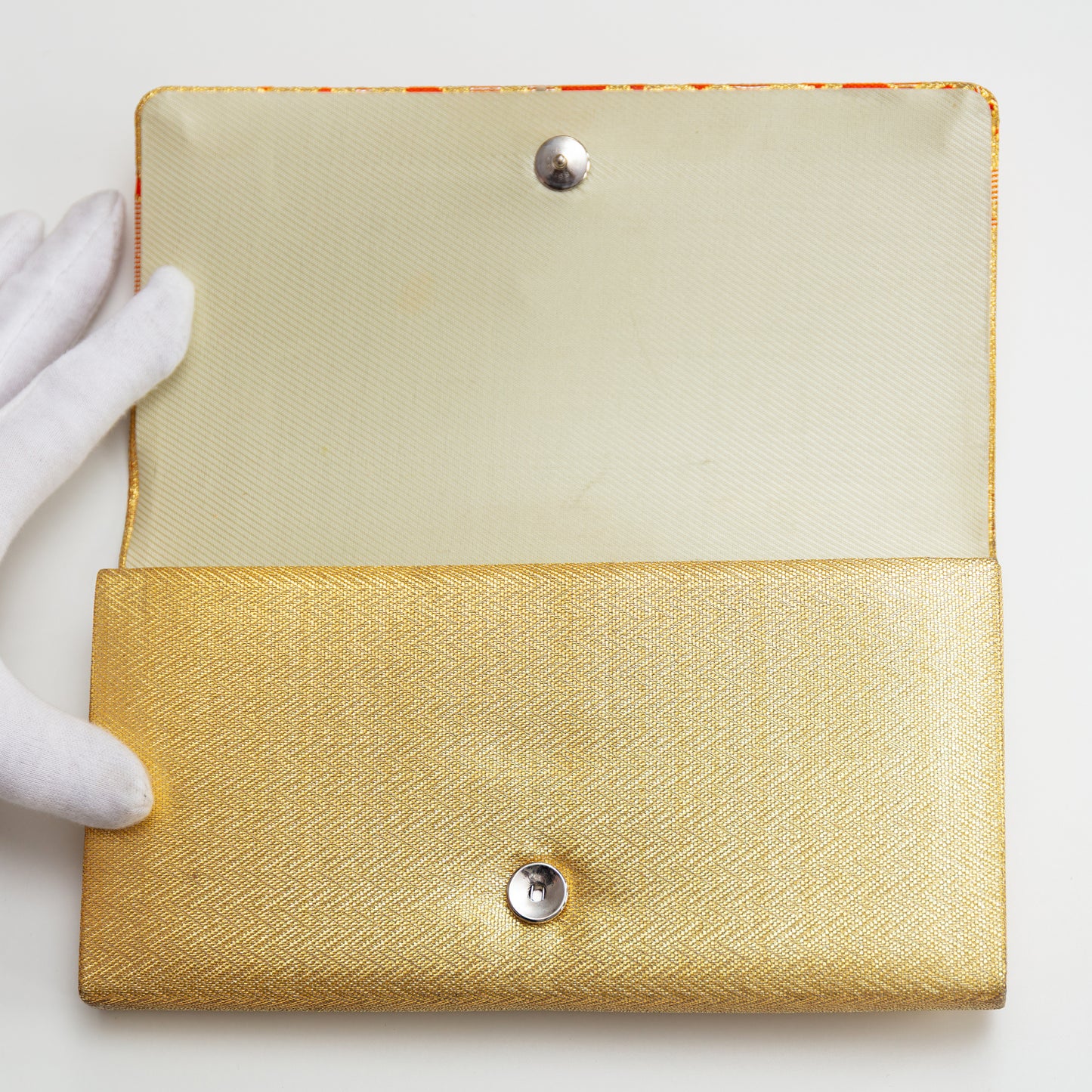 Gold clutch with textured surface and silver buttons held by a white-gloved hand on a light background