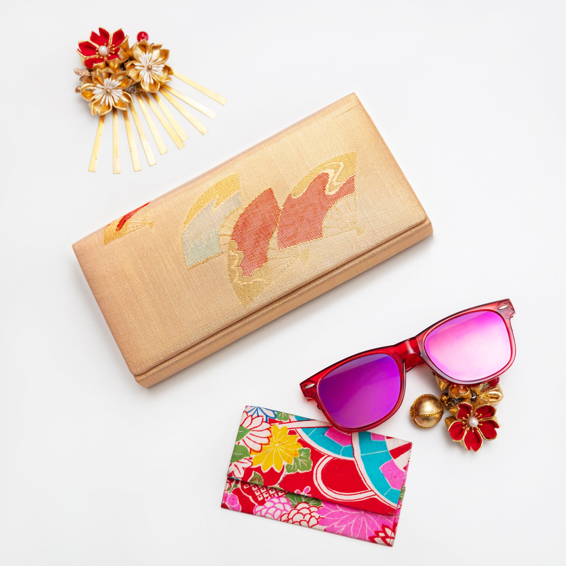 Metallic gold clutch with Japanese fans design, red sunglasses with pink lenses, colorful pouch, and decorative hairpin on a white background.