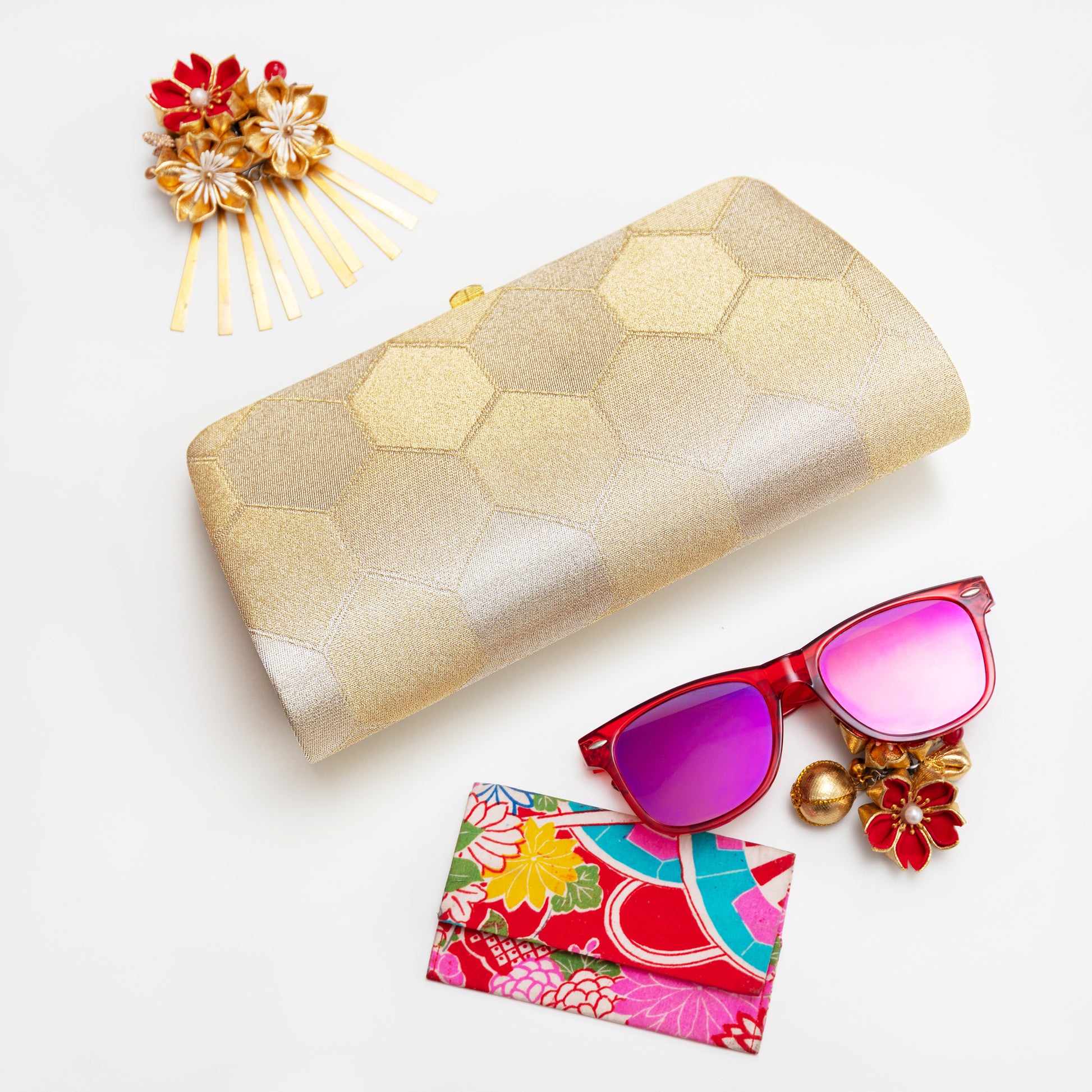 Gold hexagonal patterned clutch bag with red sunglasses and floral card on a light background