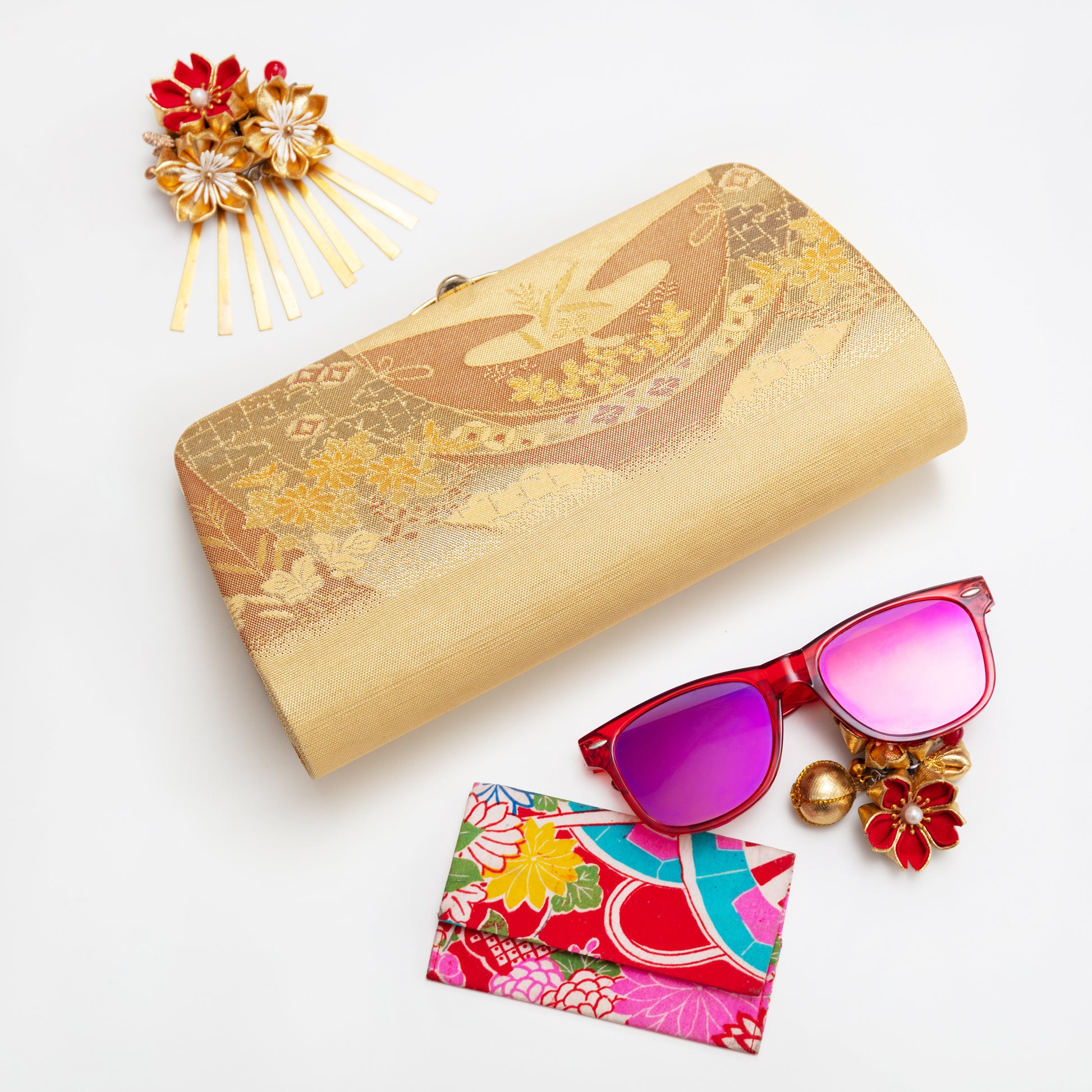 Gold clutch, red sunglasses with pink lenses, floral card holder, and decorative hairpin on a white background
