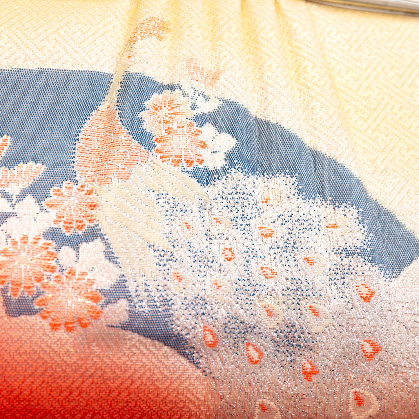 Close-up of the silk fabric front of the clutch bag with peacock and floral patterns and a gradient color scheme from peach to pale yellow /cream.