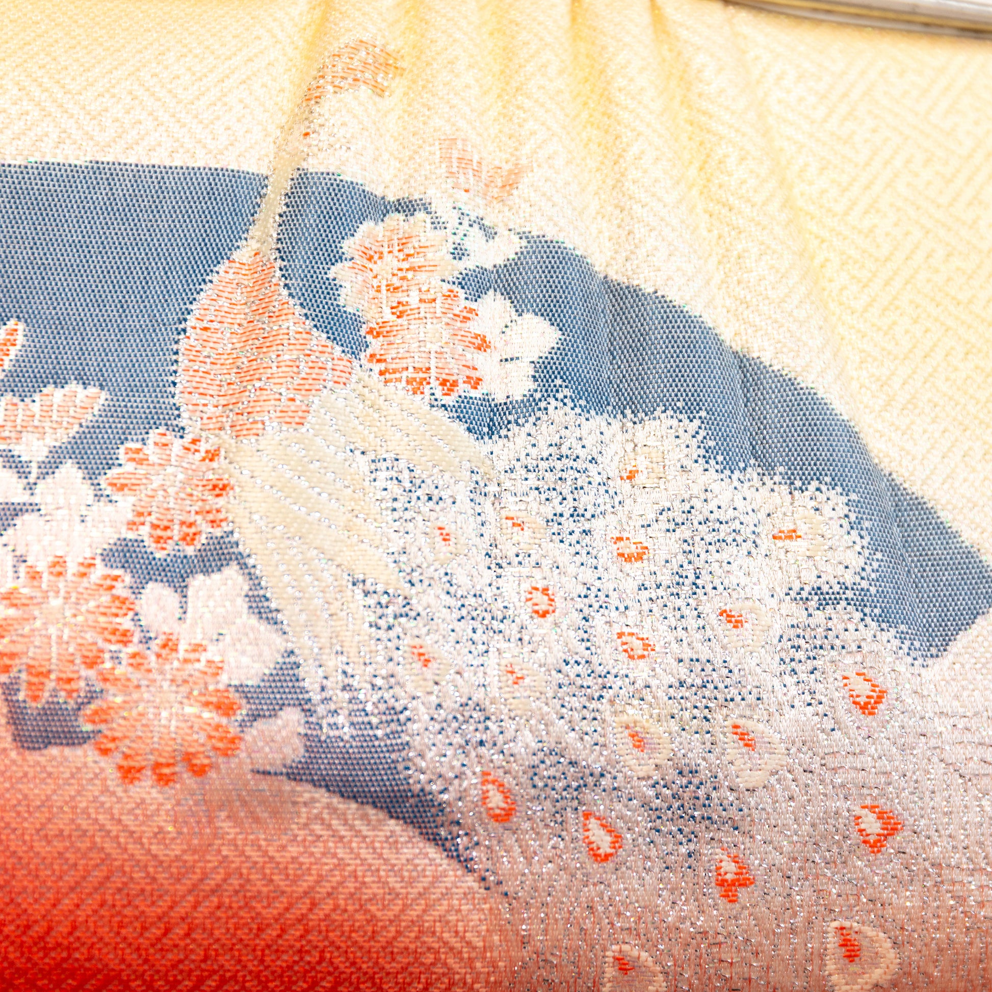 Close-up of the silk fabric front of the clutch bag with peacock and floral patterns and a gradient color scheme from peach to pale yellow /cream.