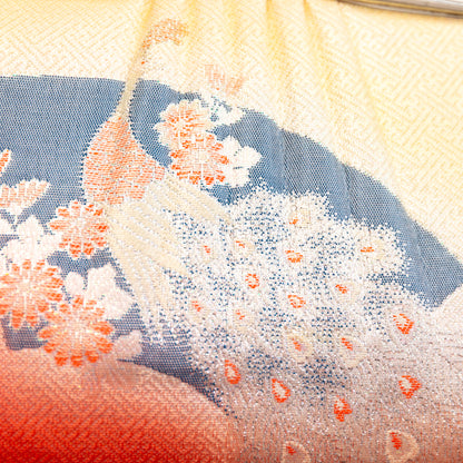 Close-up of the silk fabric front of the clutch bag with peacock and floral patterns and a gradient color scheme from peach to pale yellow /cream.