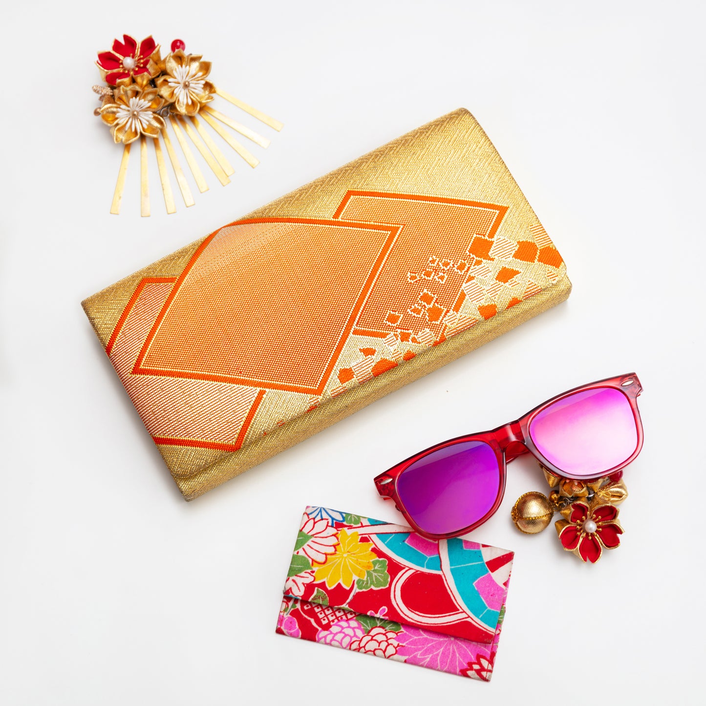 Gold clutch bag with orange diamond pattern on a white background with colourful accessories.