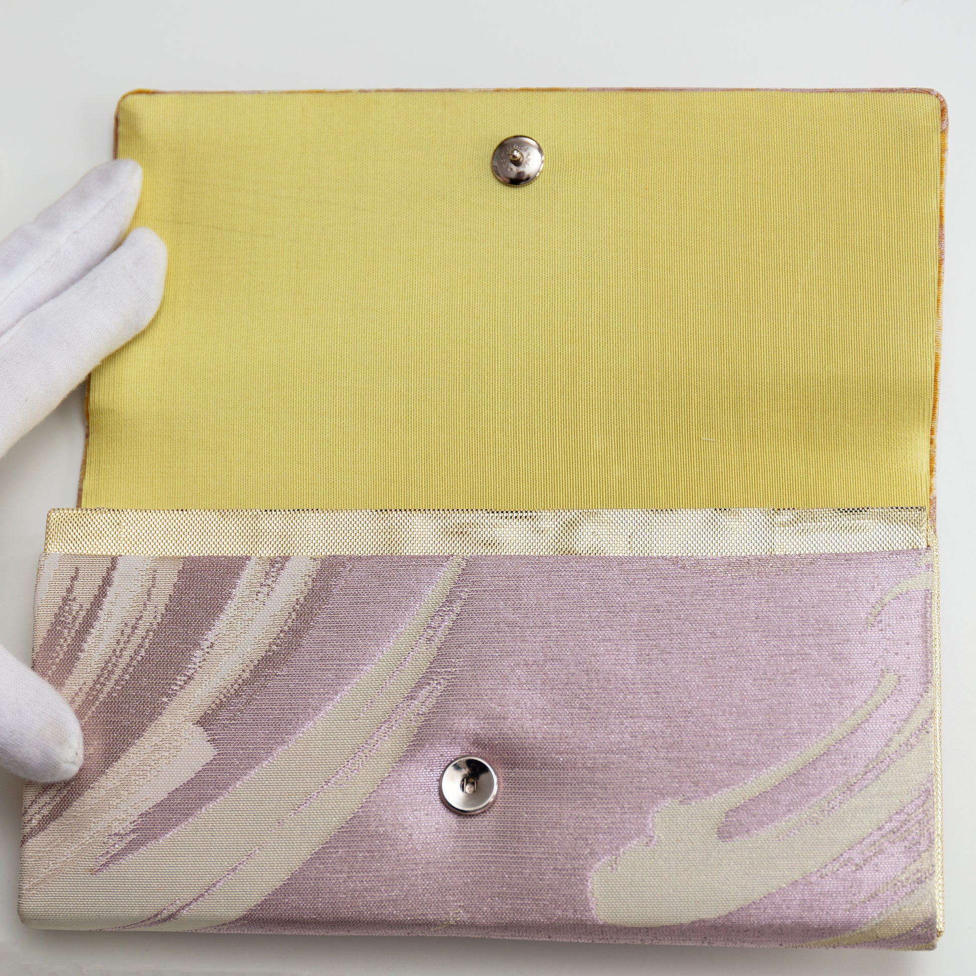 Lilac metallic clutch bag held open by a gloved hand showing the buttery cream lining silk and metal fastener.
