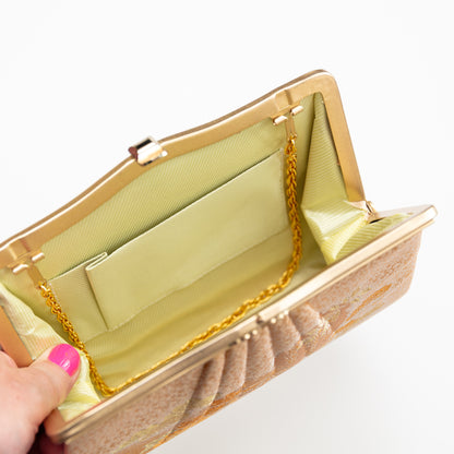 Gold clutch bag with a chain strap held by a hand on a white background. Strap can be tucked inside with convertible hinges.