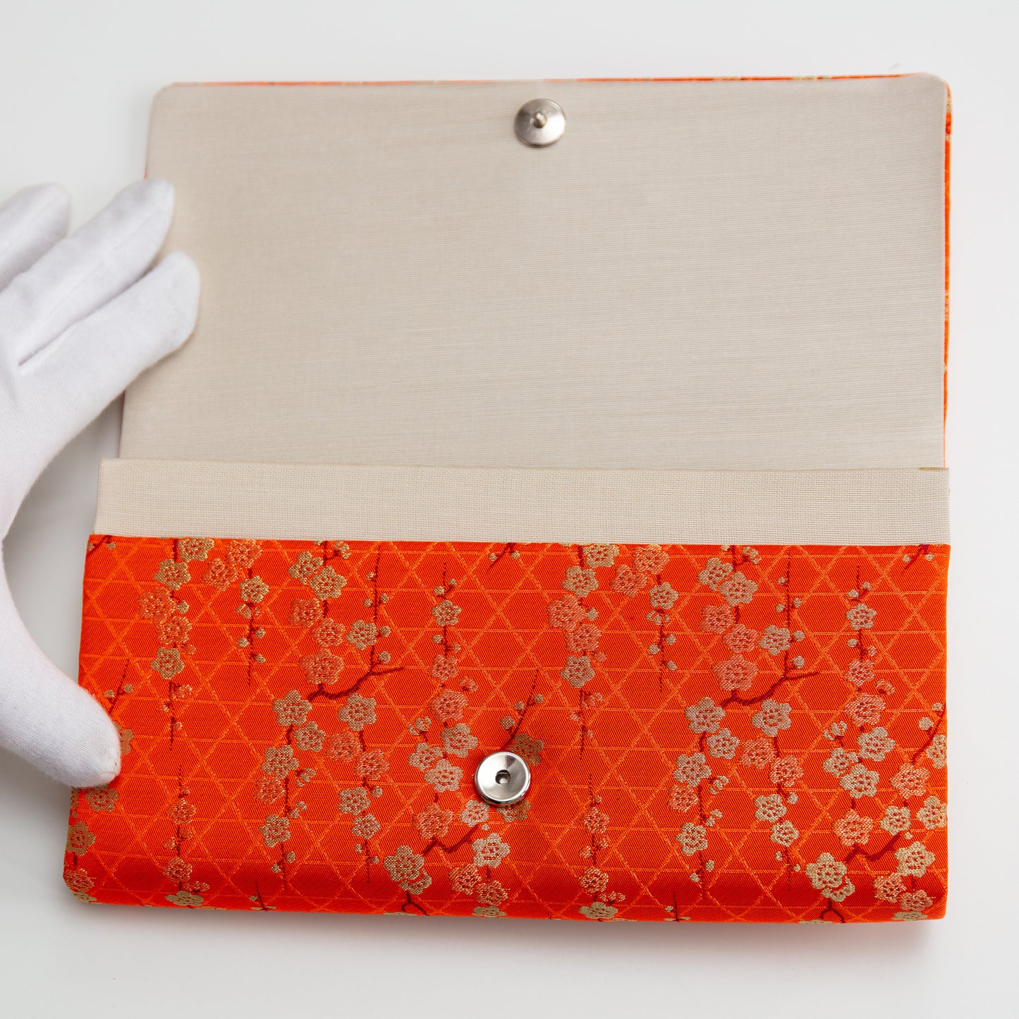 Floral-patterned orange and gold clutch bag held open by white gloved hand to show beige silk interior lining and silver fastener on a white background