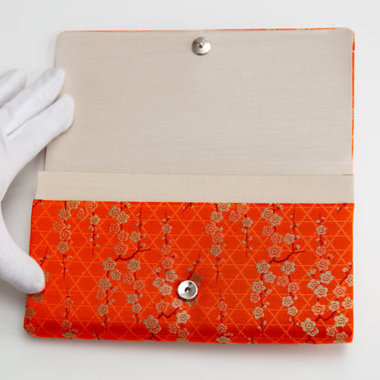 Floral-patterned orange and gold clutch bag held open by white gloved hand to show beige silk interior lining and silver fastener on a white background